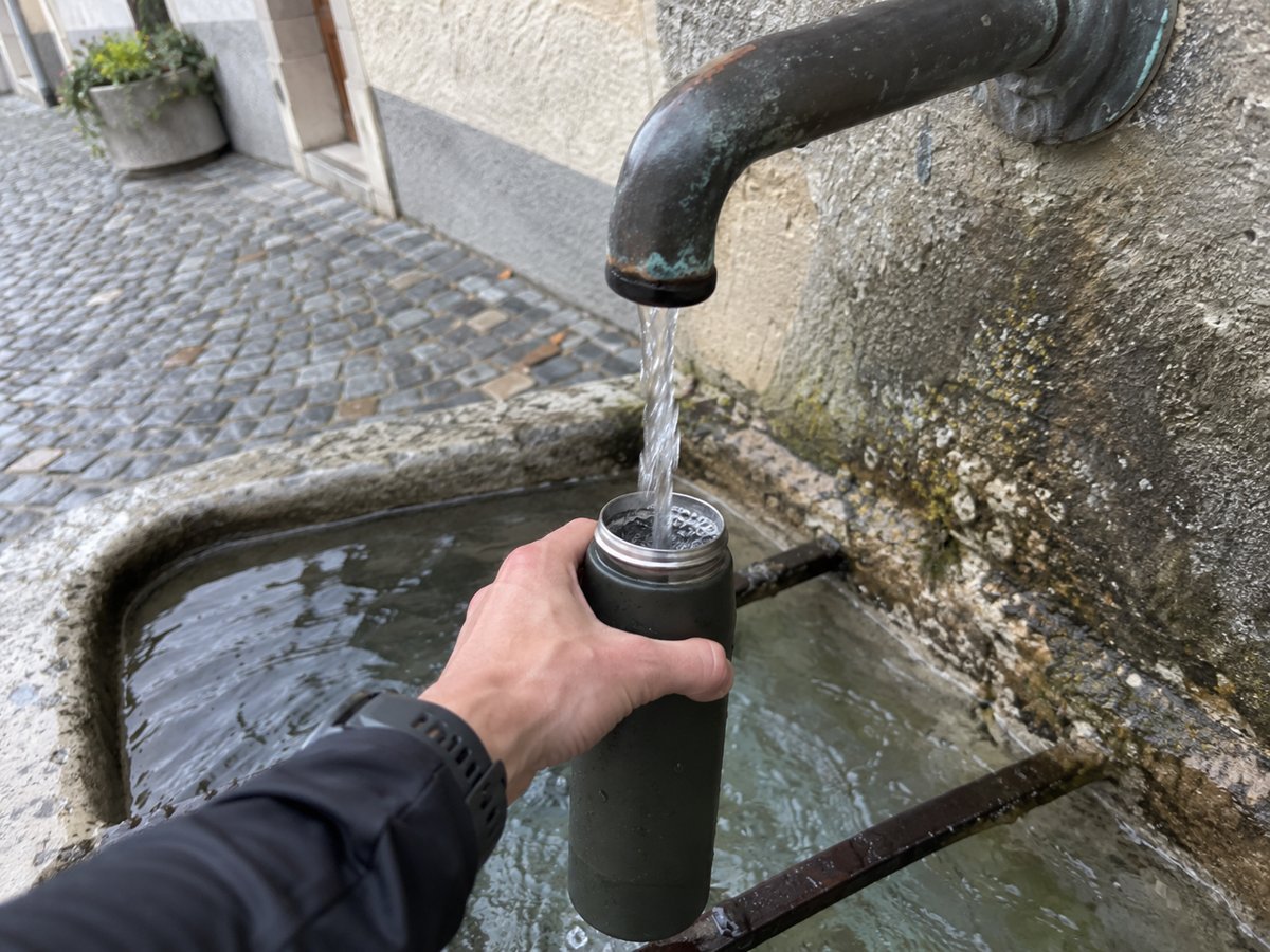 Eine Hand hält eine geöffnete Trinkflasche unter den Wasserstrahl eines alten Brunnens.