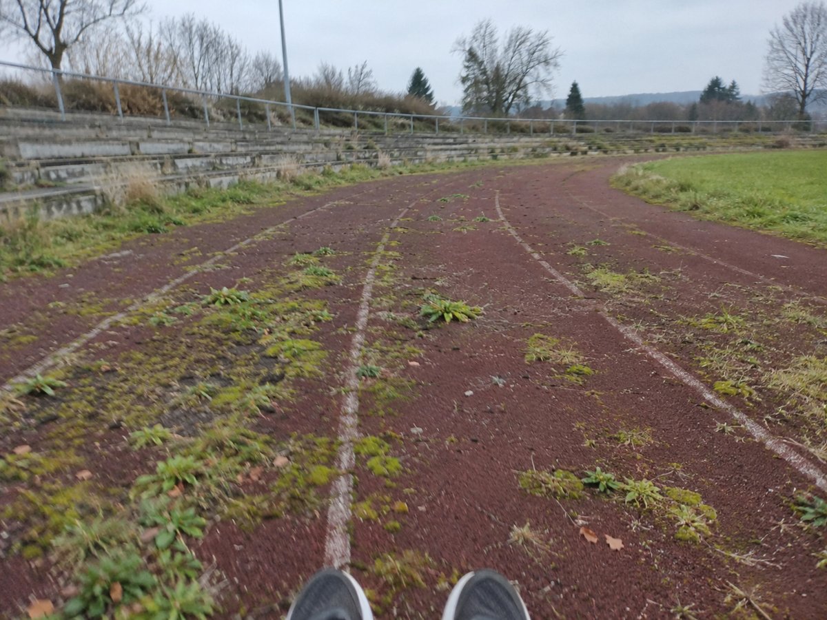 Alte rotbraune Aschenbahn mit Unkraut und Moos in den Bahnen, dahinter niedrige Betonstufen eines kleinen Stadions.