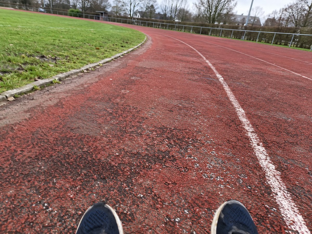 Abgenutzte rote Laufbahn mit weißer Linie und Schuhspitzen am unteren Bildrand.