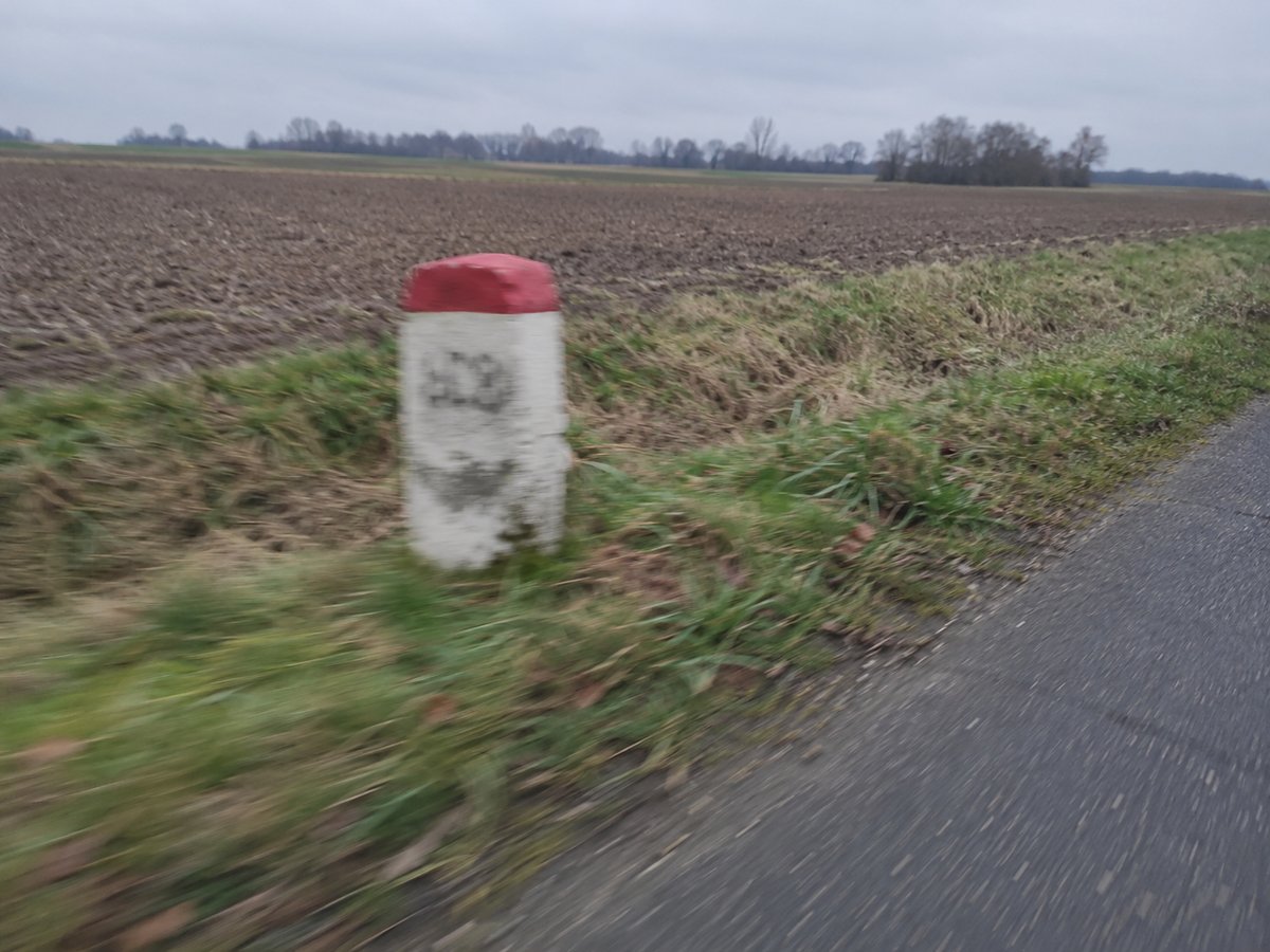 Kleiner weißer Kilometerstein mit roter Kappe am Rand einer Landstraße, verwackelt aufgenommen.