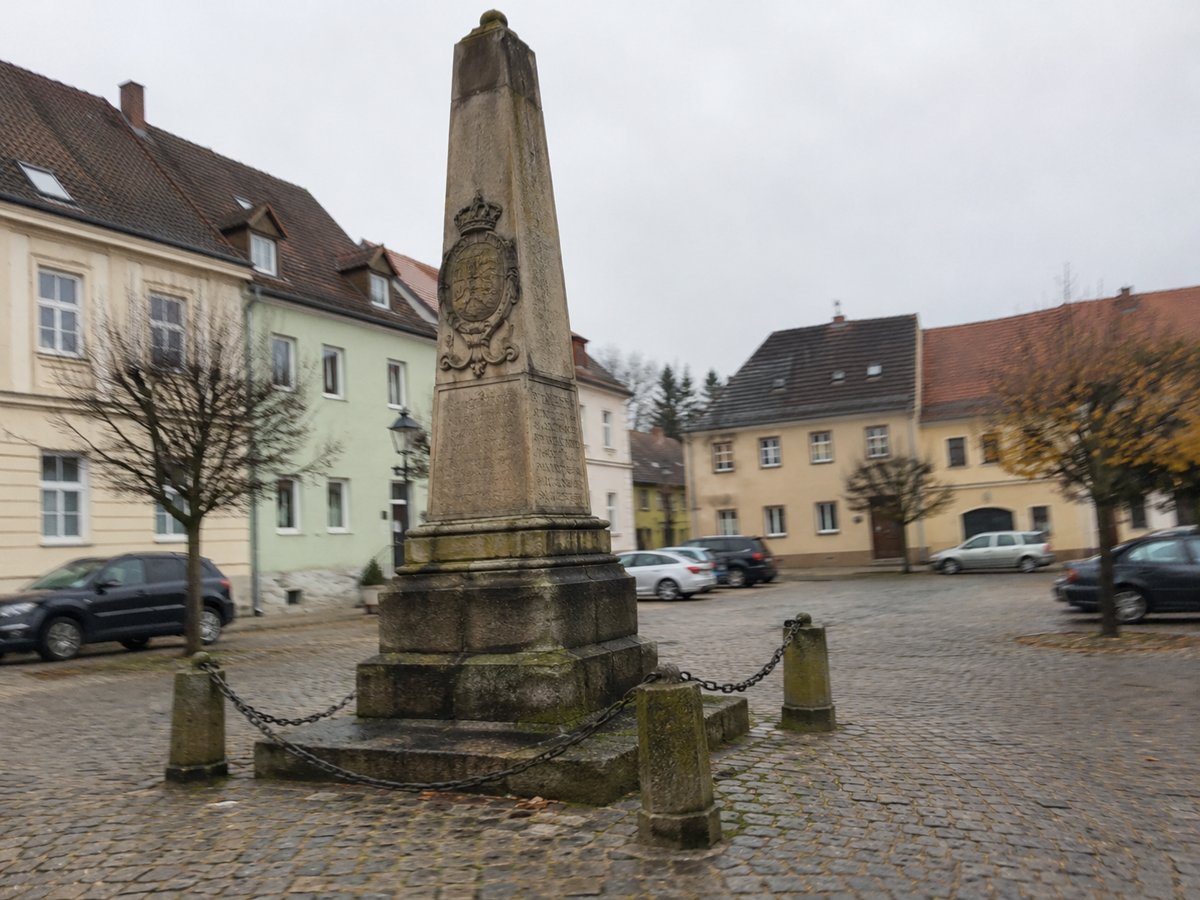 Distanzsäule auf einem gepflasterten Kleinstadtplatz, dahinter unscharfe geparkte Autos und alte Häuser.