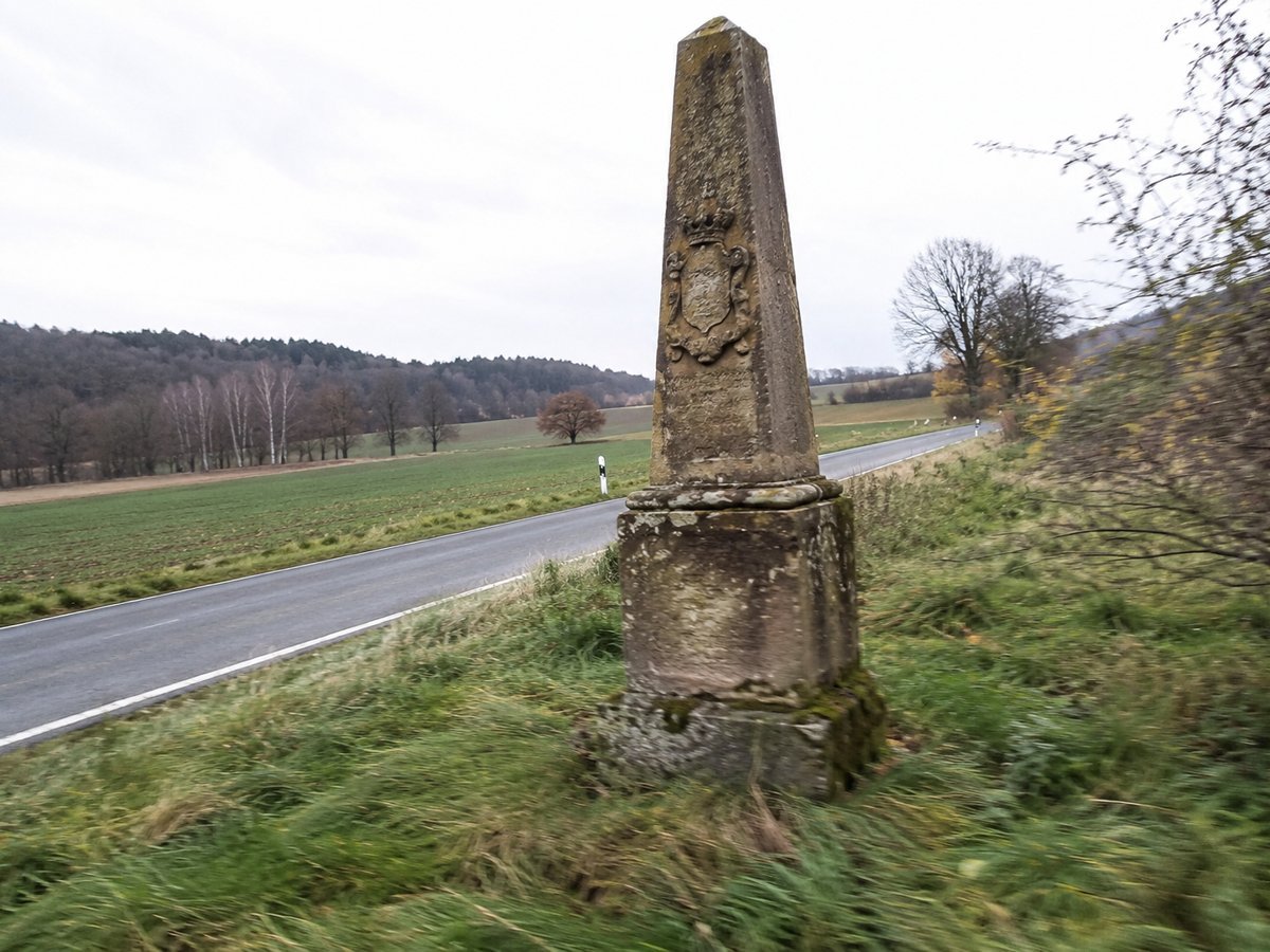 Verwitterte Postmeilensäule aus Sandstein am grasbewachsenen Rand einer Landstraße.